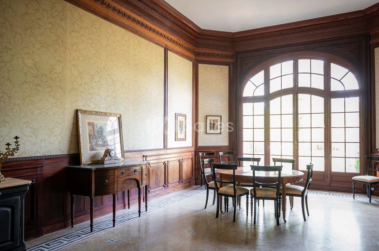 Salle à manger lumineuse avec table, chaises en bois, buffet ancien, grandes fenêtres et murs ornés de boiseries.