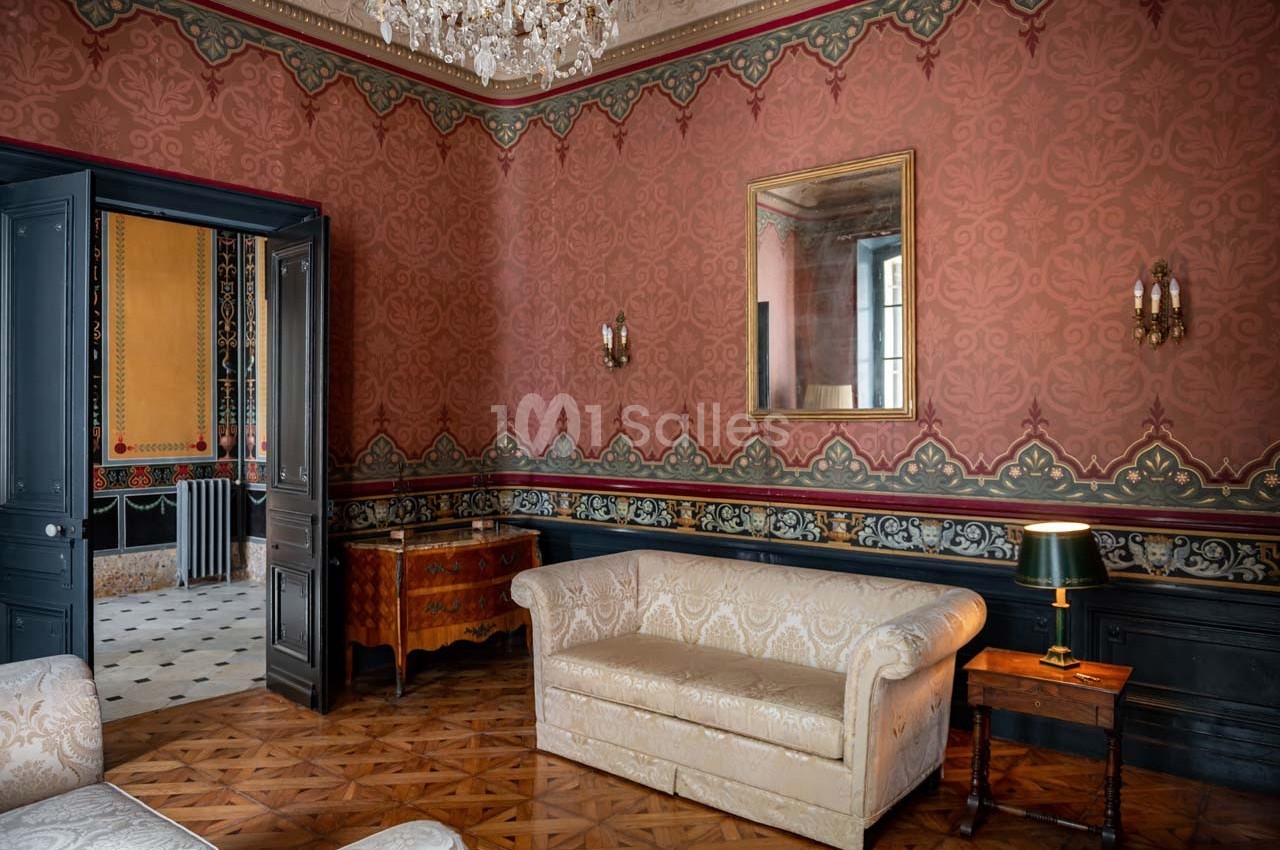Salon ancien avec canapé beige, murs ornés de motifs rouges, moulures décoratives, lustre et miroir doré.