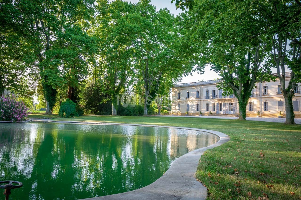 Étang bordé d'herbe et d'arbres, avec un grand bâtiment clair en arrière-plan sous un ciel dégagé.