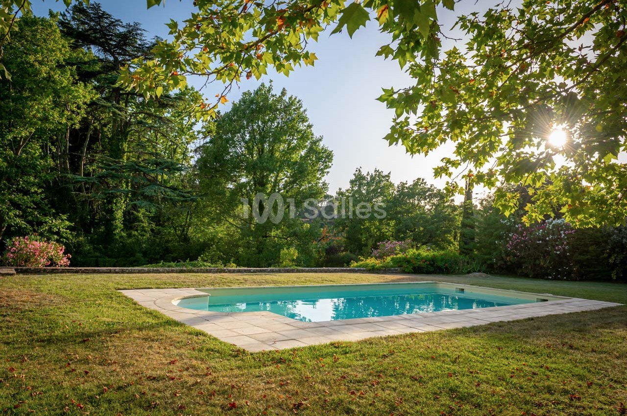 Piscine extérieure entourée de pelouse et d'arbres, sous un ciel dégagé avec le soleil filtrant à travers les branches.