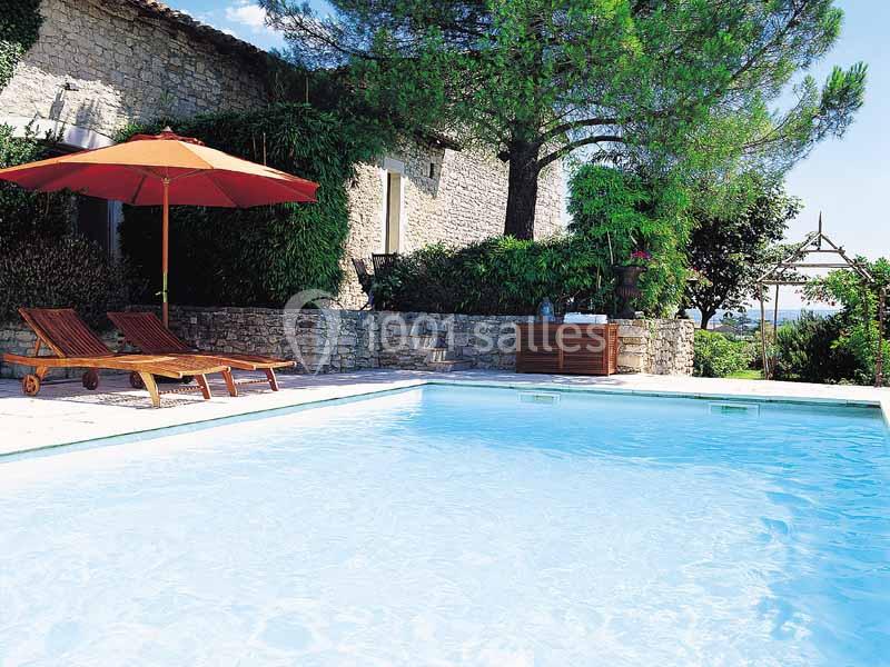 Piscine extérieure entourée de chaises longues, parasol rouge, végétation et mur en pierre sous un ciel dégagé.
