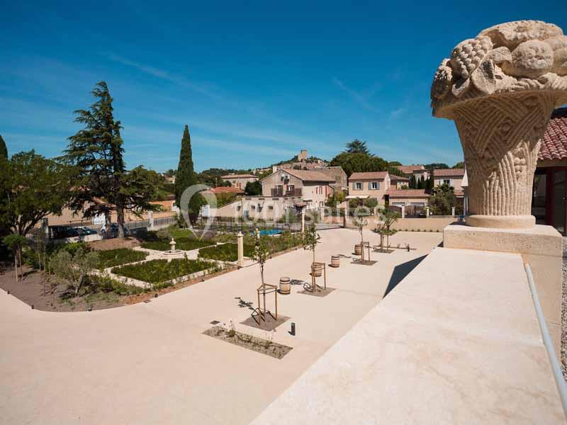 Vue d'une place ensoleillée avec des jardins, des arbres, des maisons et une sculpture en pierre au premier plan.