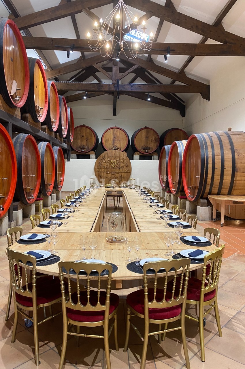 Salle de dégustation avec une longue table en bois entourée de chaises dorées, entourée de grandes barriques en bois.
