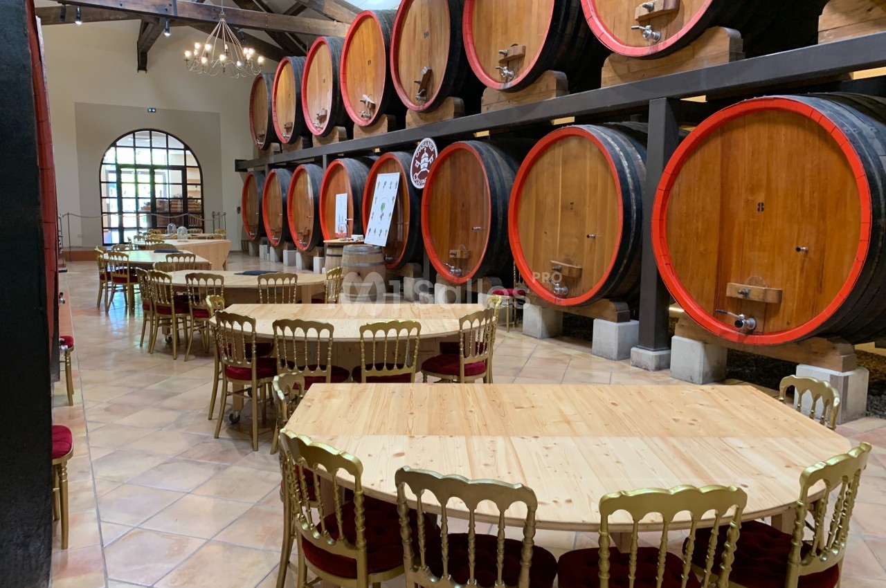 Salle de dégustation avec tables en bois et chaises dorées, entourée de grandes barriques de vin alignées.