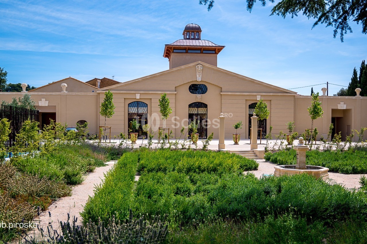 Bâtiment beige avec une coupole centrale, entouré d'un jardin verdoyant et de chemins en gravier.