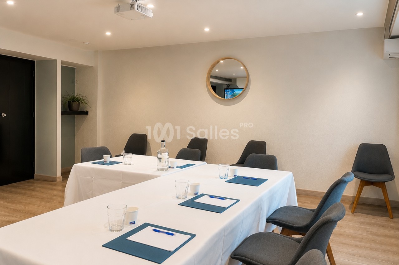 Salle de réunion lumineuse avec tables blanches, chaises grises, blocs-notes et un miroir rond au mur.