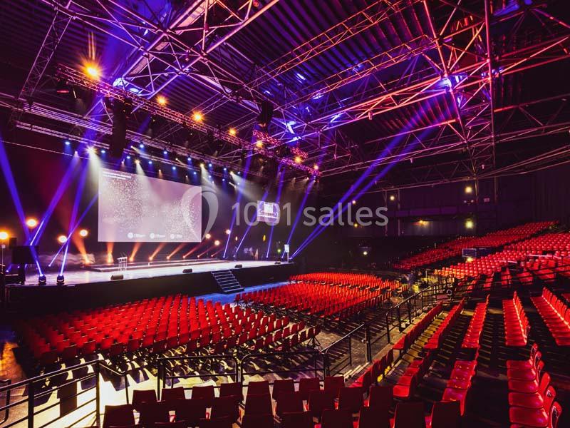 Salle de spectacle vide avec sièges rouges, scène éclairée par des projecteurs et structure métallique visible au plafond.