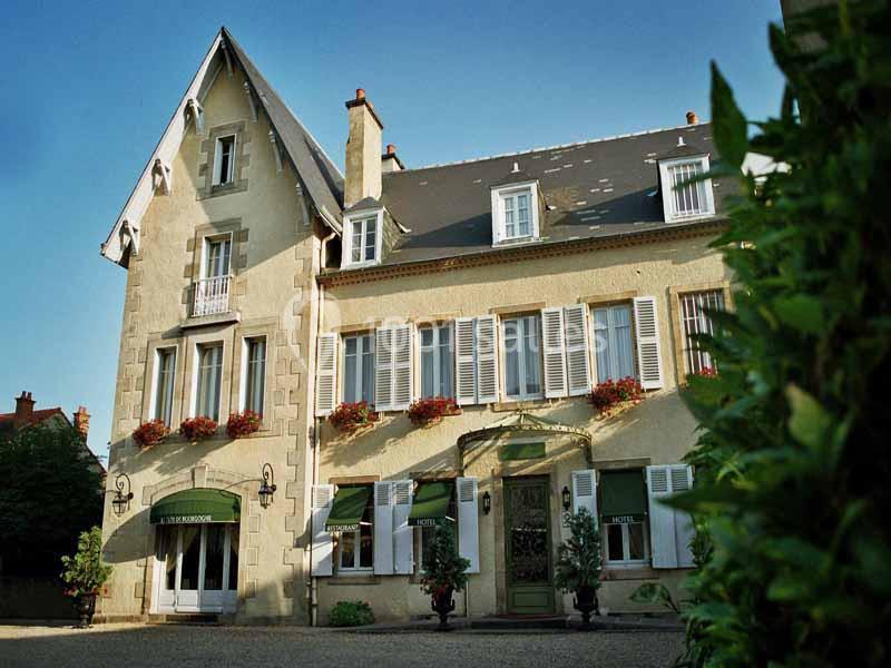 Façade d'un hôtel de style traditionnel avec volets blancs, jardinières fleuries et entrée principale entourée de verdure.