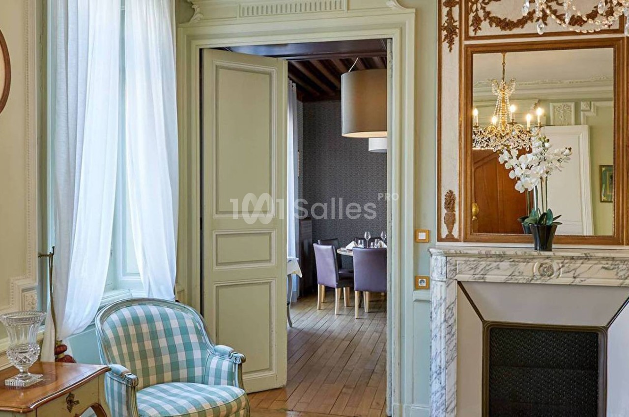 Salon élégant avec cheminée en marbre, fauteuil à carreaux, et vue sur une salle à manger lumineuse en arrière-plan.