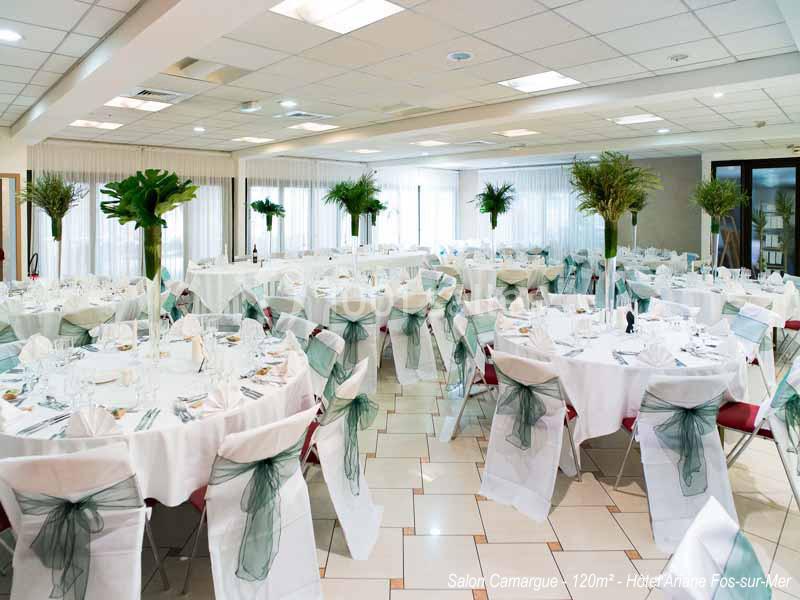 Salle de réception lumineuse avec tables rondes dressées, chaises décorées de nœuds verts et centres de table fleuris.