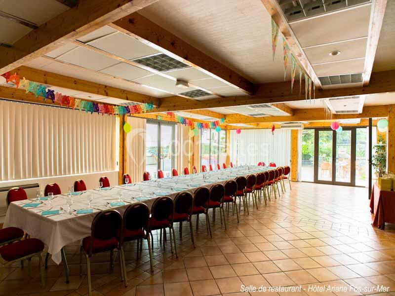 Grande salle lumineuse avec une longue table dressée pour un repas, décorée de guirlandes colorées et de ballons.