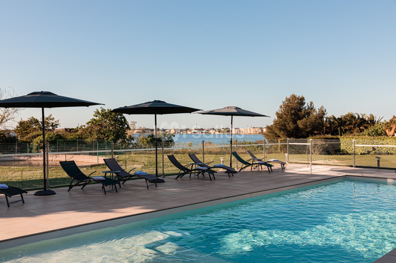 Piscine extérieure avec transats et parasols, entourée d'un espace vert et offrant une vue sur un plan d'eau.