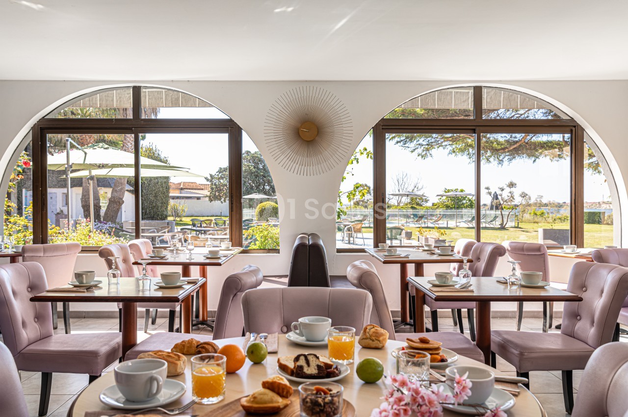 Salle lumineuse avec tables dressées pour le petit-déjeuner, vue sur un jardin et terrasse en arrière-plan.