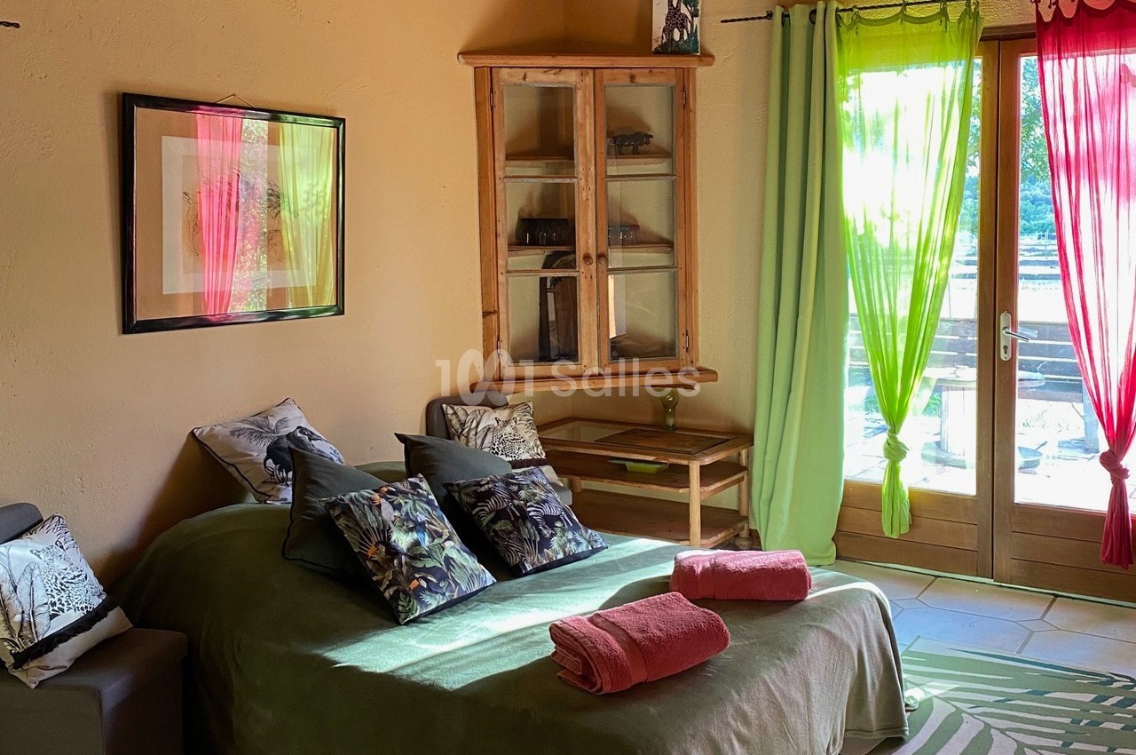 Chambre lumineuse avec lit recouvert de coussins et serviettes, armoire en bois, rideaux colorés et porte-fenêtre donnant…