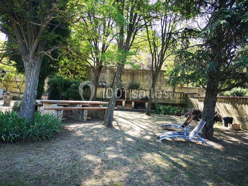 Espace extérieur ombragé avec des arbres, des chaises longues, une table en bois et un mur en pierre en arrière-plan.