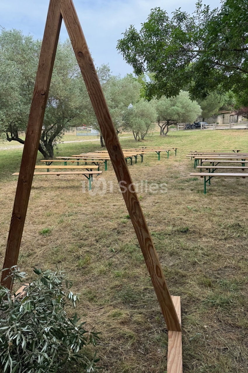 Structure en bois triangulaire devant des bancs disposés en plein air sur une pelouse entourée d'arbres.