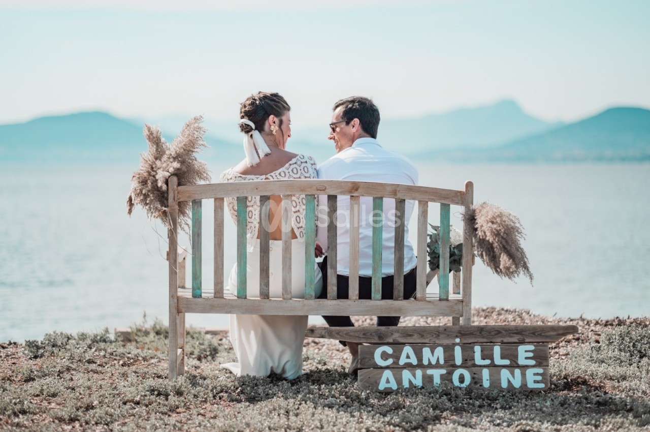 Un couple assis sur un banc en bois près d'un lac, entouré de pampas, avec une pancarte portant leurs prénoms.