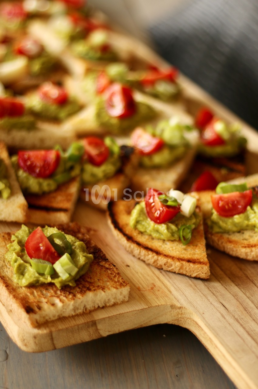 Tranches de pain grillé garnies de guacamole, de tomates cerises et de ciboulette sur une planche en bois.