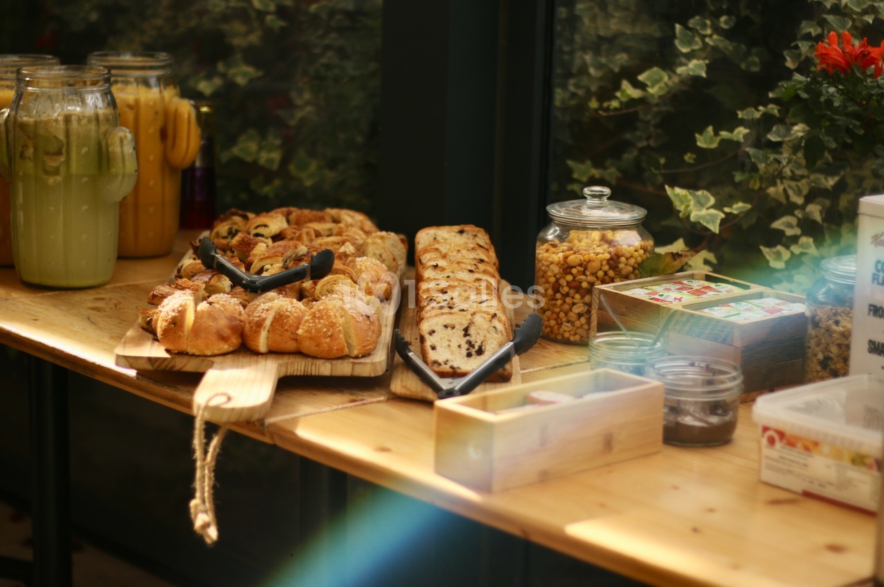 Table en bois avec viennoiseries, pain, bocaux de céréales et jus dans un cadre lumineux entouré de verdure.