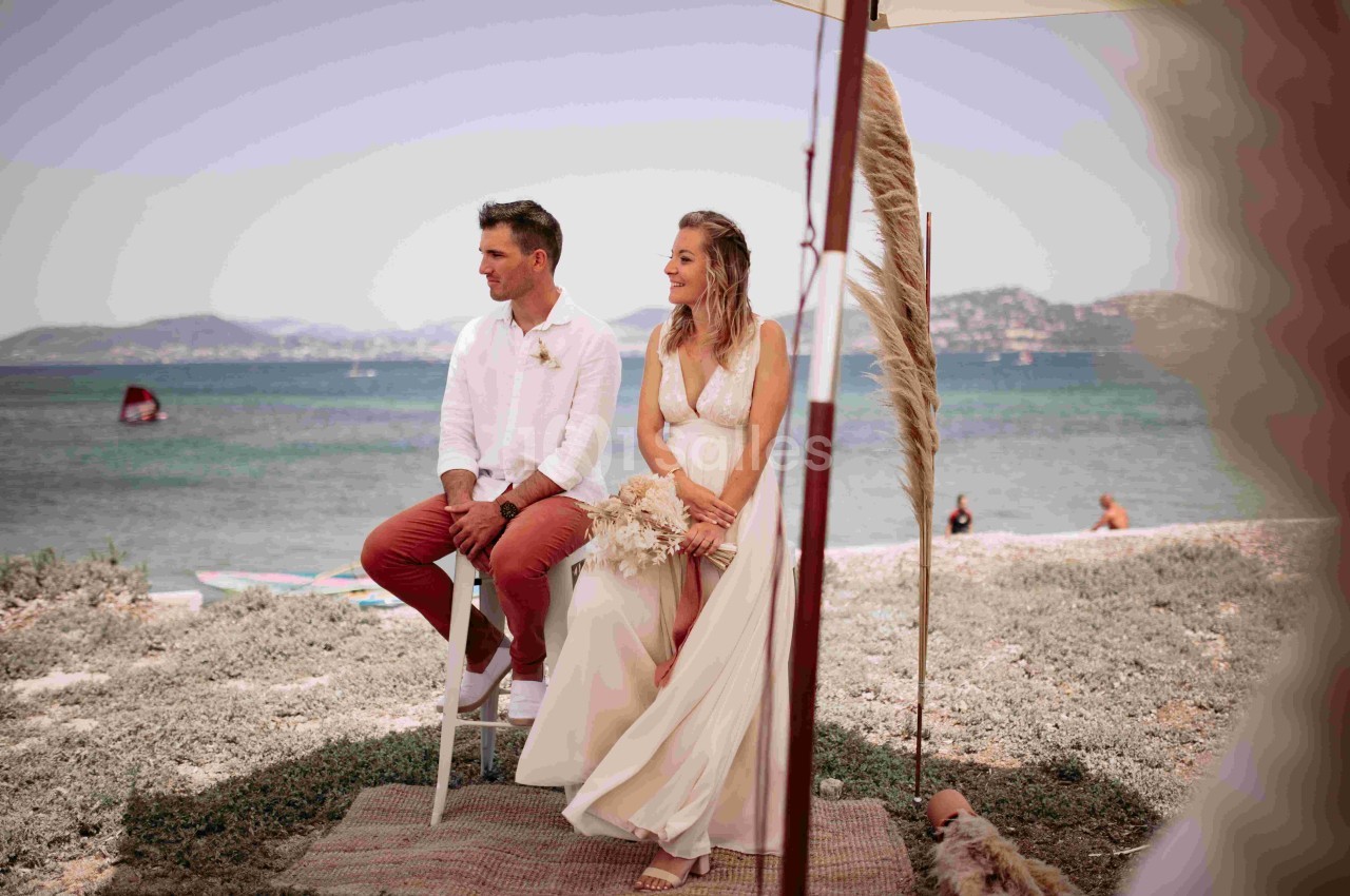 Un couple assis sous un parasol près de la mer, entouré d'herbe et d'éléments décoratifs naturels.