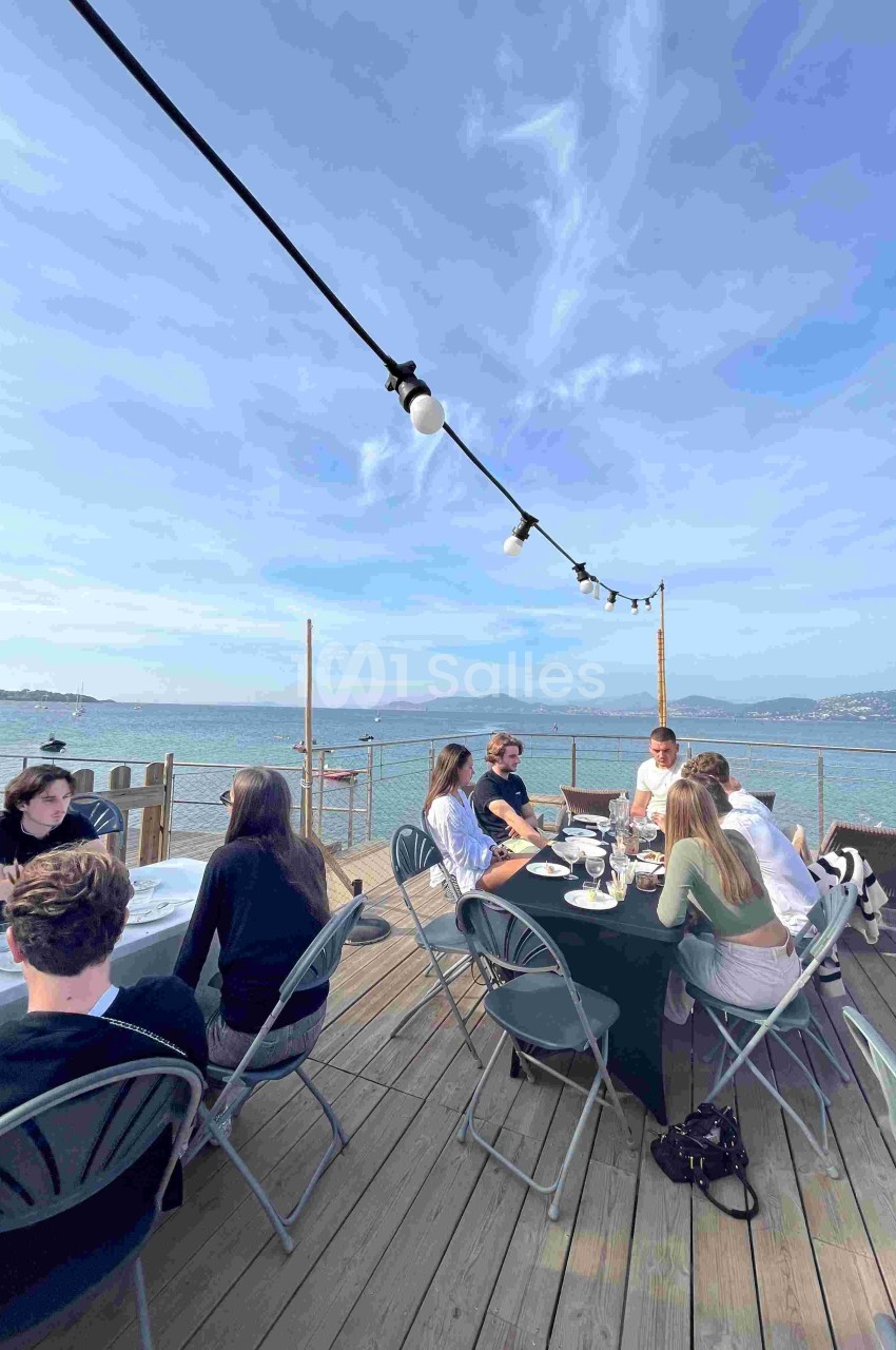 Des personnes assises autour de tables sur une terrasse en bois, avec vue sur la mer et un ciel dégagé.