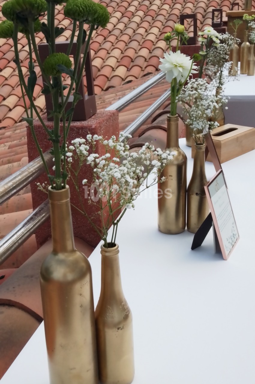 Bouteilles dorées recyclées servant de vases avec des fleurs blanches et vertes, disposées sur une table en extérieur.