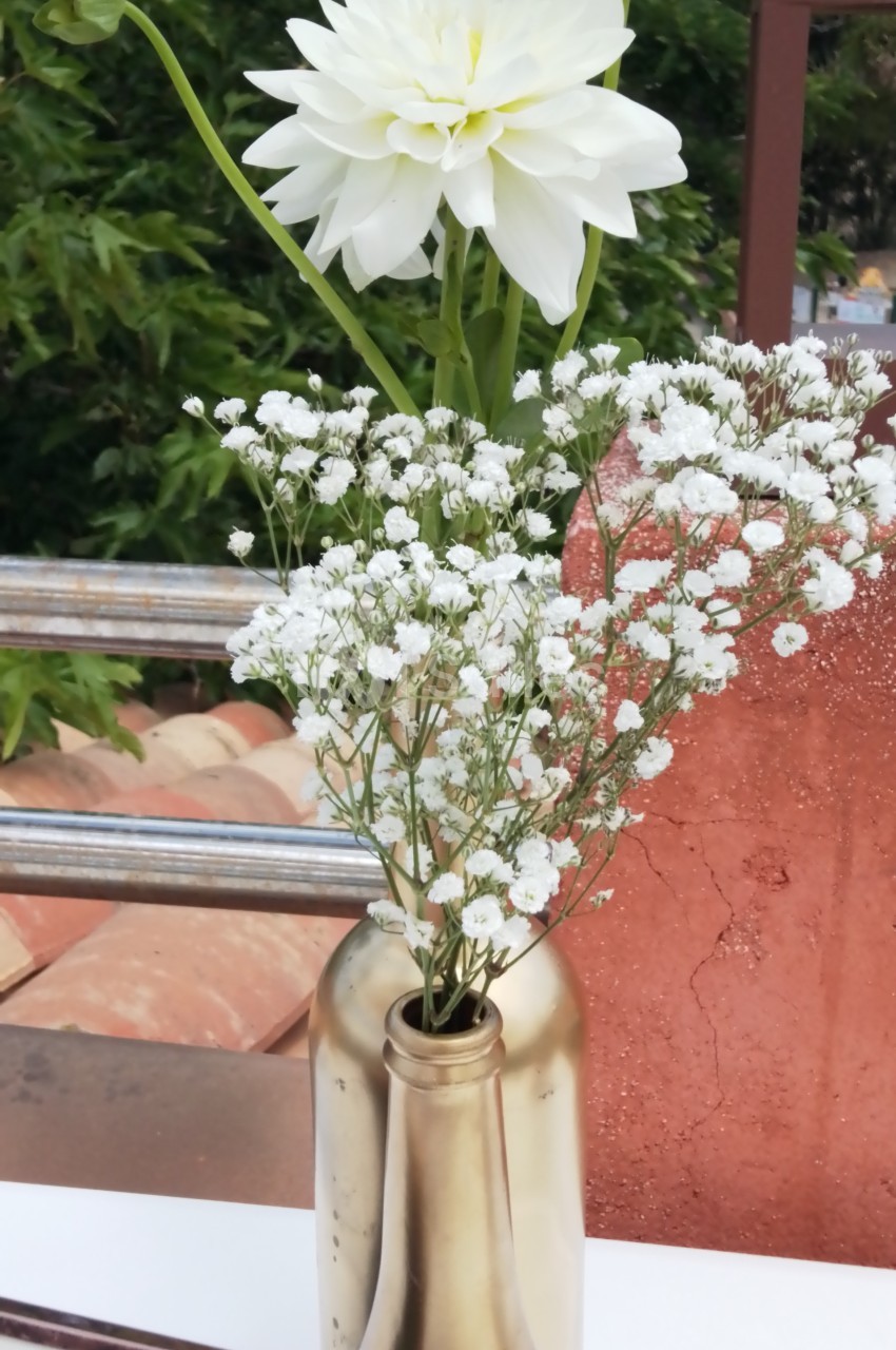 Fleur blanche et gypsophile disposées dans un vase doré, en extérieur avec un fond de végétation et de briques.