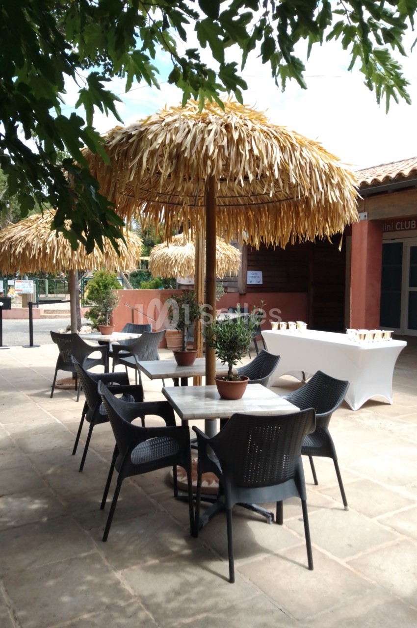 Terrasse avec tables rondes, chaises noires et parasols en paille, entourée de verdure et d'un bâtiment en arrière-plan.