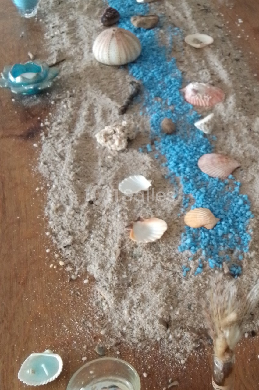 Décoration de table avec sable, coquillages, galets et un chemin de gravier bleu évoquant un cours d'eau.