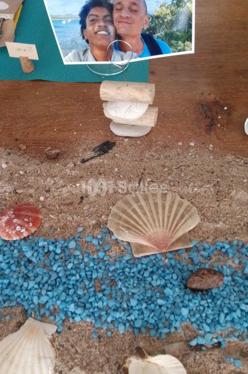 Coquillages disposés sur du sable avec des gravillons bleus, photo de deux personnes souriantes en arrière-plan.