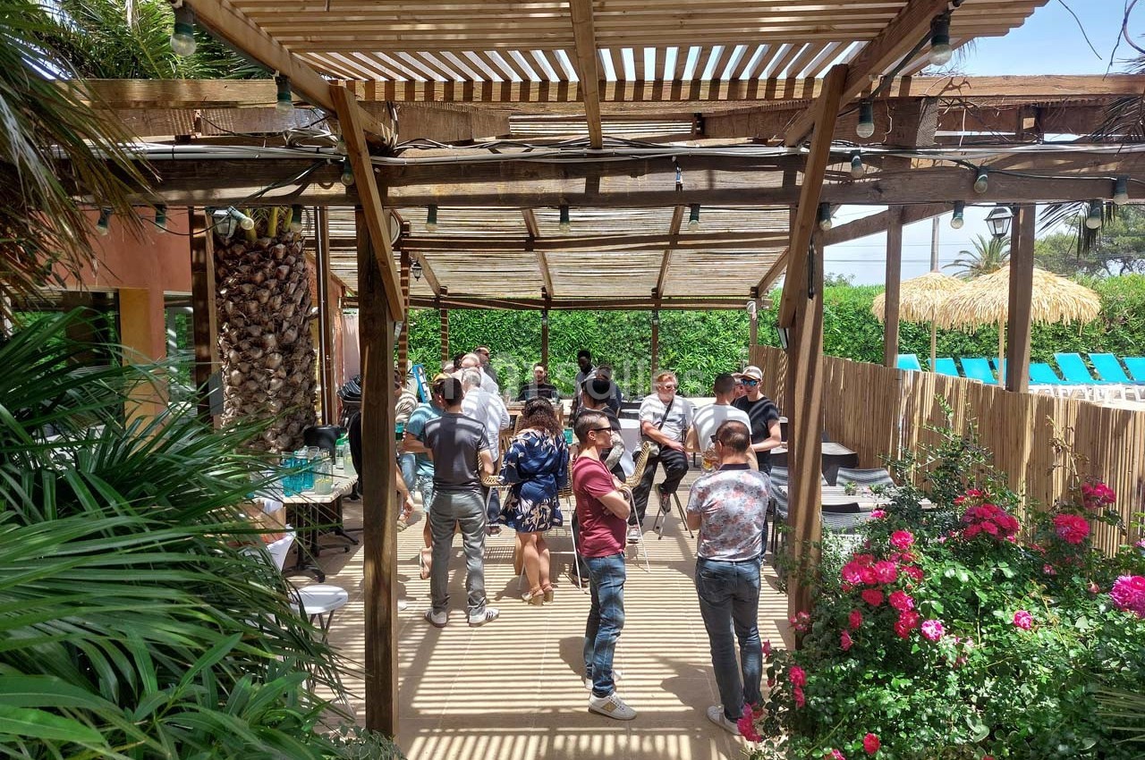 Groupe de personnes discutant sous une pergola en bois, entourée de végétation et de fleurs colorées.