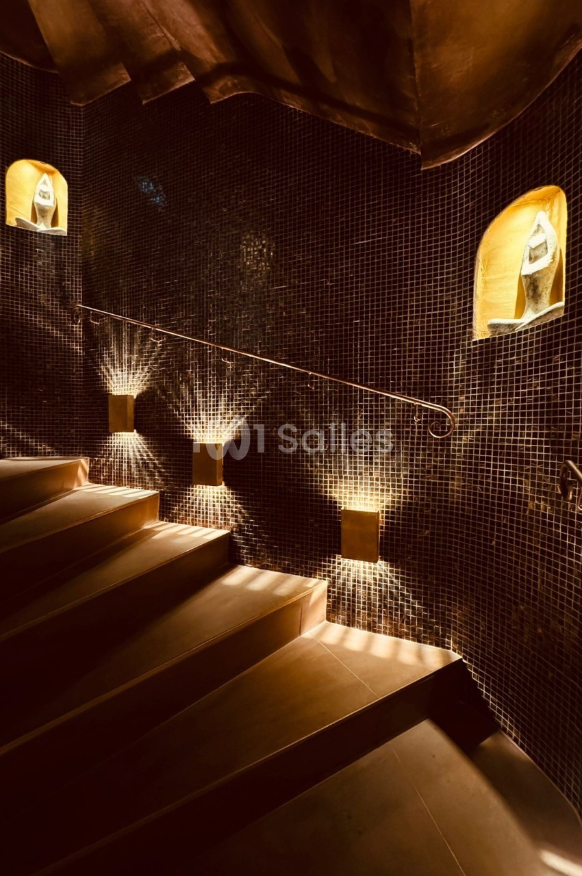 Escalier en colimaçon avec des murs en mosaïque sombre, éclairé par des niches lumineuses contenant des objets décoratifs.