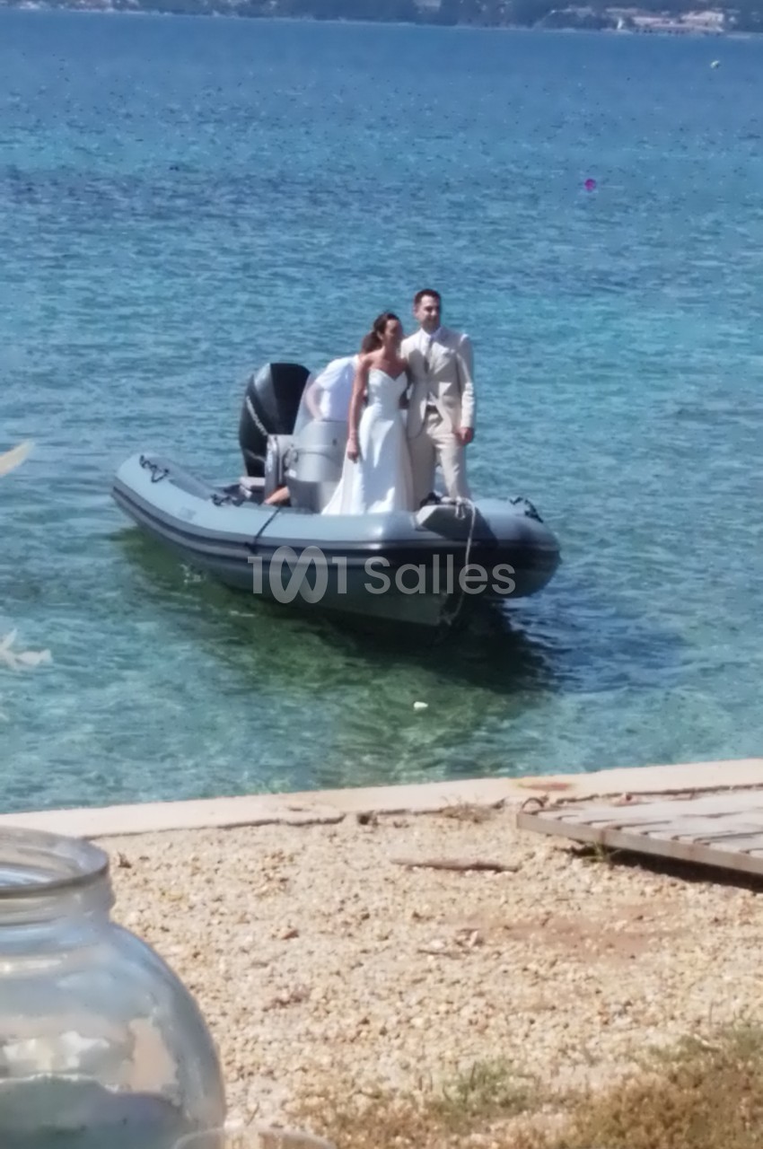 Un couple en tenue de mariage se tient debout sur un bateau pneumatique amarré près d'une plage.