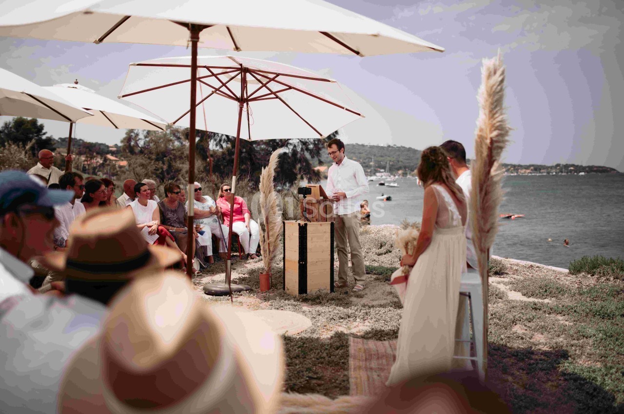 Un homme fait un discours devant un groupe de personnes assises sous des parasols lors d'un événement en plein air.