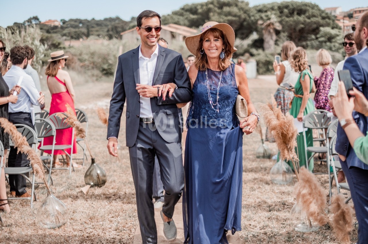 Un homme en costume et une femme en robe bleue avancent ensemble lors d'une cérémonie en extérieur.