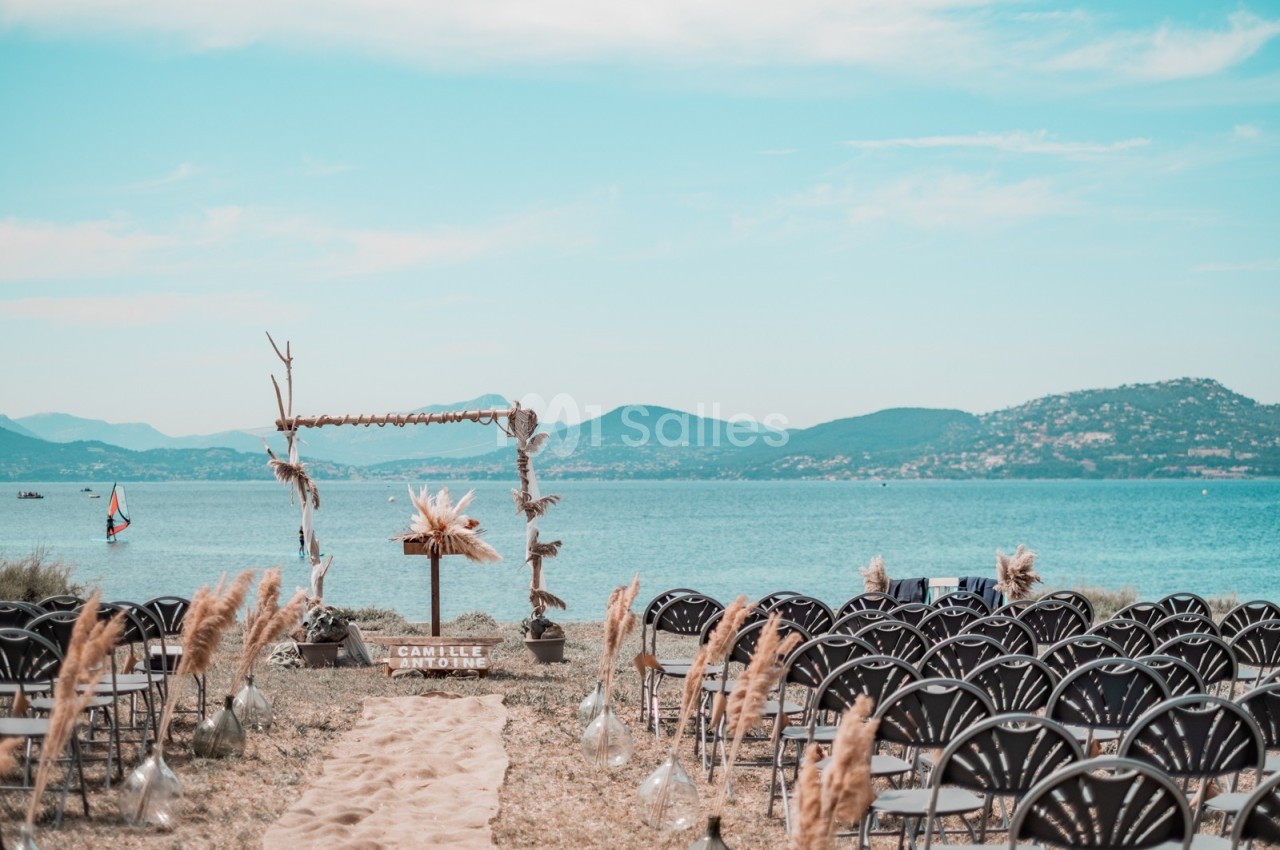 Chaises disposées face à une arche décorée en bord de mer, avec vue sur des collines et un ciel dégagé.