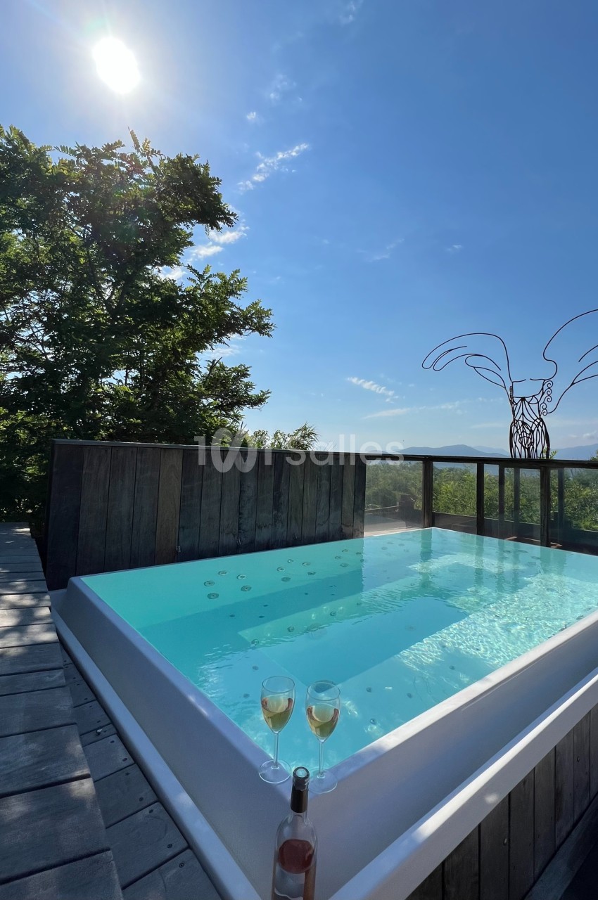 Bain à remous extérieur sur une terrasse en bois, entouré de verdure, avec deux verres de vin et vue dégagée sous un ciel…