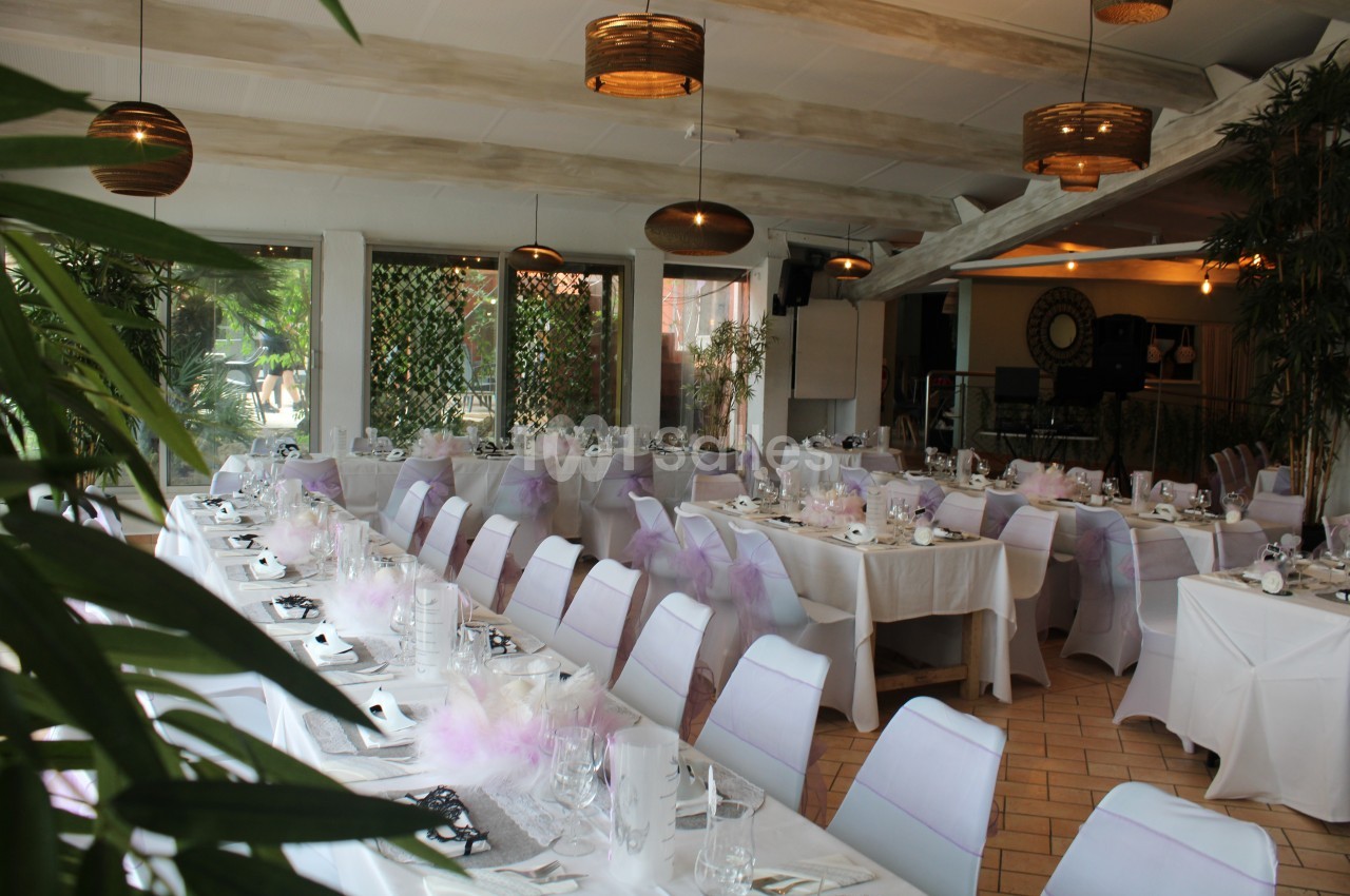 Salle de réception lumineuse avec tables dressées, nappes blanches et décorations mauves, entourée de plantes.