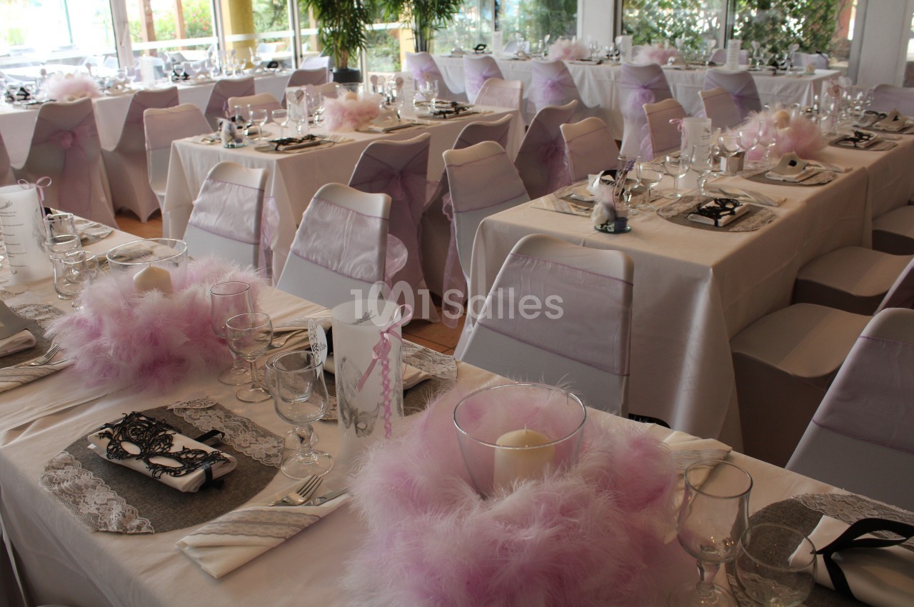 Salle de réception décorée avec des tables dressées, nappes blanches, centres de table roses et chaises ornées de rubans.
