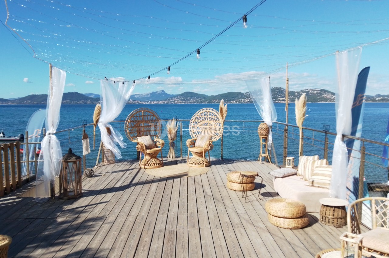 Terrasse en bois avec mobilier en rotin, vue sur la mer, montagnes en arrière-plan et ciel dégagé.