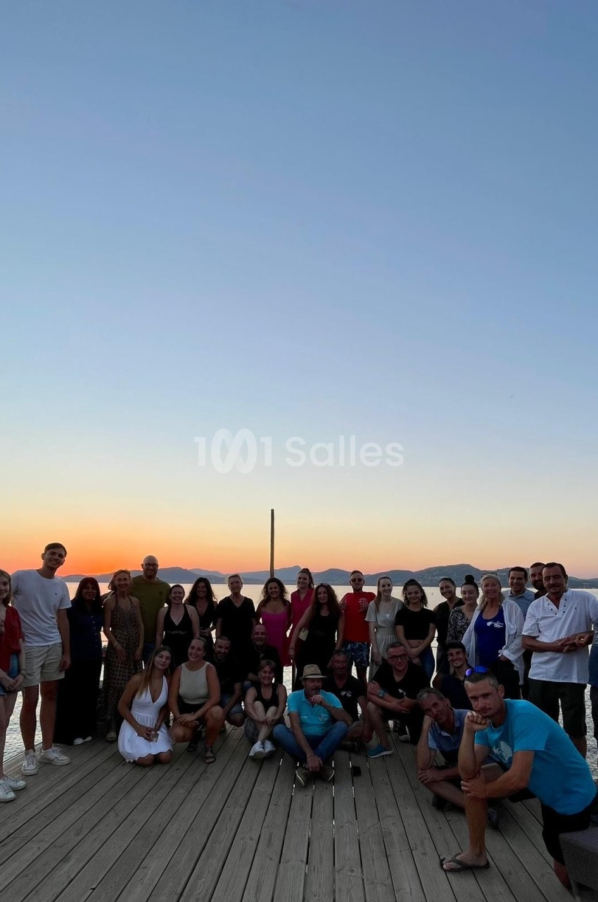 Groupe de personnes posant sur une terrasse en bois au coucher du soleil avec une vue sur la mer et des montagnes.
