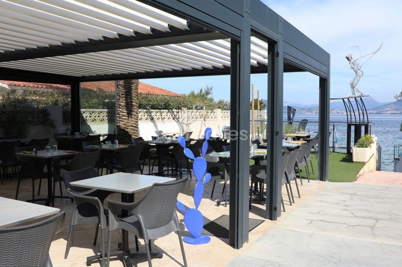 Terrasse d'un restaurant avec pergola, tables et chaises, vue sur la mer et une sculpture moderne en arrière-plan.