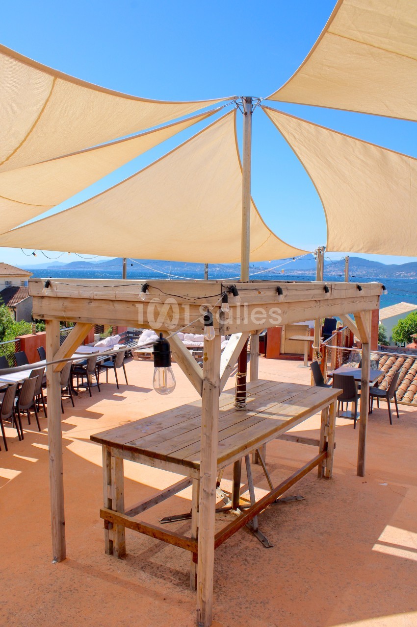 Table en bois sous des voiles d'ombrage beige sur une terrasse avec vue sur un paysage côtier en arrière-plan.