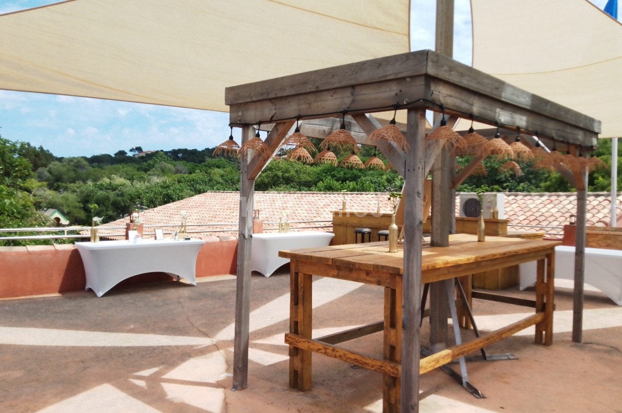 Table en bois sous une pergola avec des lampes suspendues, sur une terrasse extérieure ombragée par des voiles tendues.