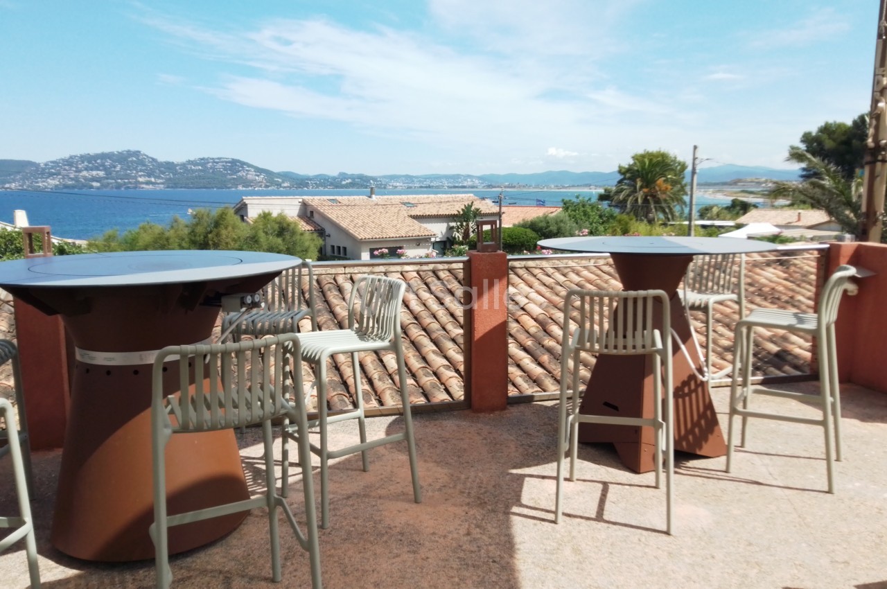 Terrasse avec tables hautes, chaises en métal et vue sur des toits en tuiles, la mer et des collines en arrière-plan.