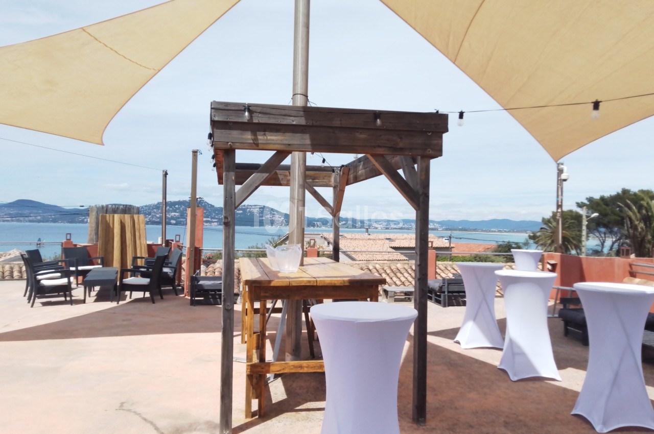 Terrasse ensoleillée avec vue sur la mer, mobilier en bois et tables hautes recouvertes de nappes blanches.