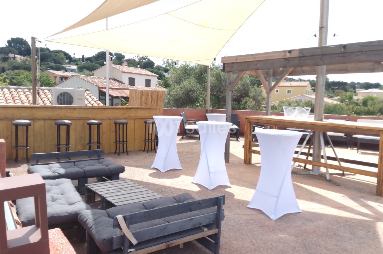 Terrasse aménagée avec canapés, tables hautes, tabourets et pergola, entourée de maisons et végétation.