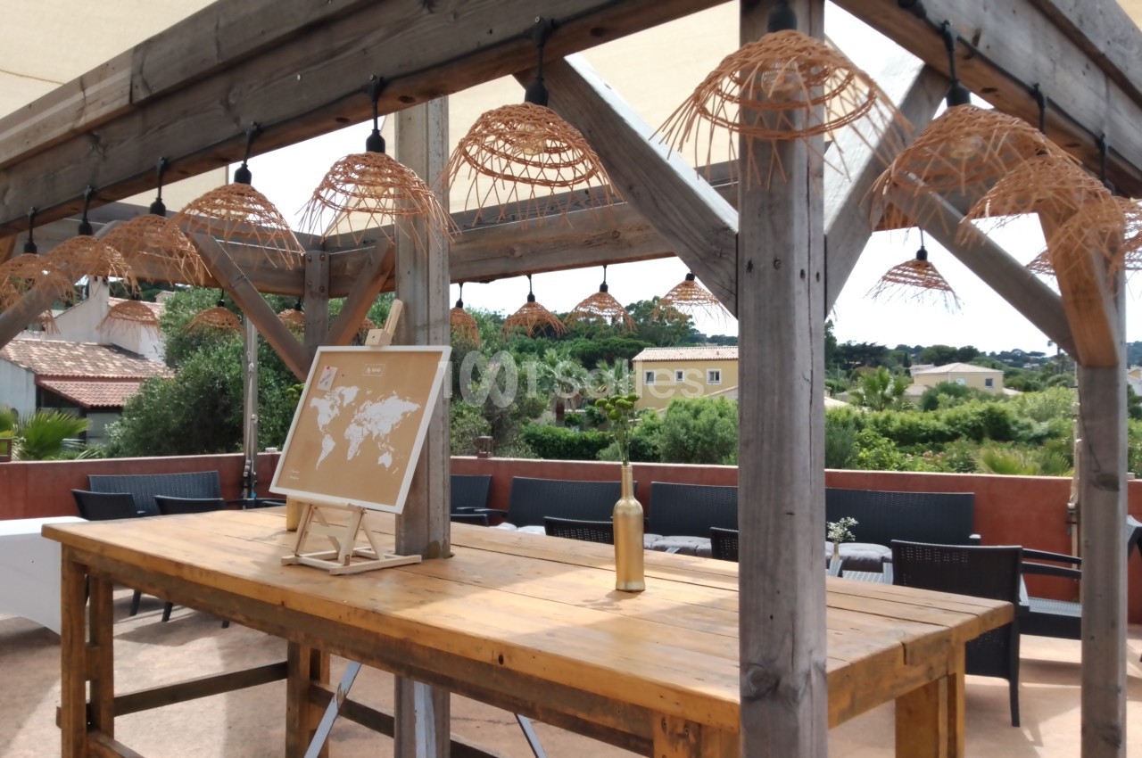 Table en bois sous une pergola avec suspensions en osier, carte du monde posée dessus, vue sur des jardins en arrière-plan.