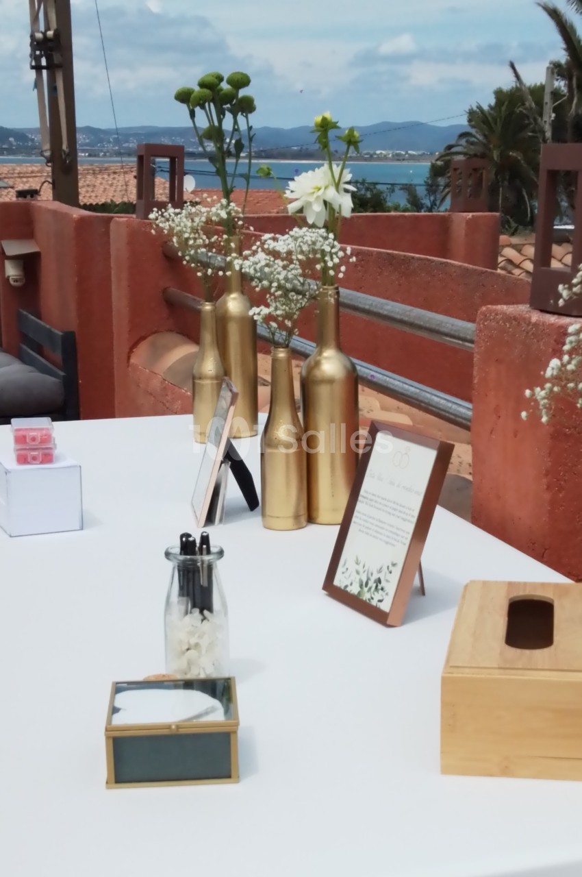 Table décorée avec des vases dorés contenant des fleurs, sur une terrasse avec vue sur la mer et des toits en arrière-plan.