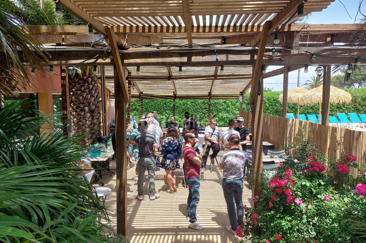 Groupe de personnes discutant sous une pergola en bois, entourée de végétation et de fleurs colorées.
